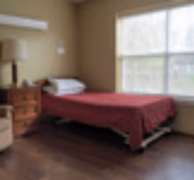 A simple bedroom with a single bed covered in a red blanket, a wooden nightstand with a lamp, and a large window letting in natural light.