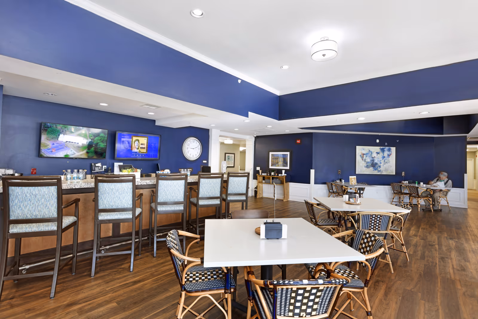 Dining area with a bar counter and high stools, multiple tables and chairs, and TVs on blue walls.