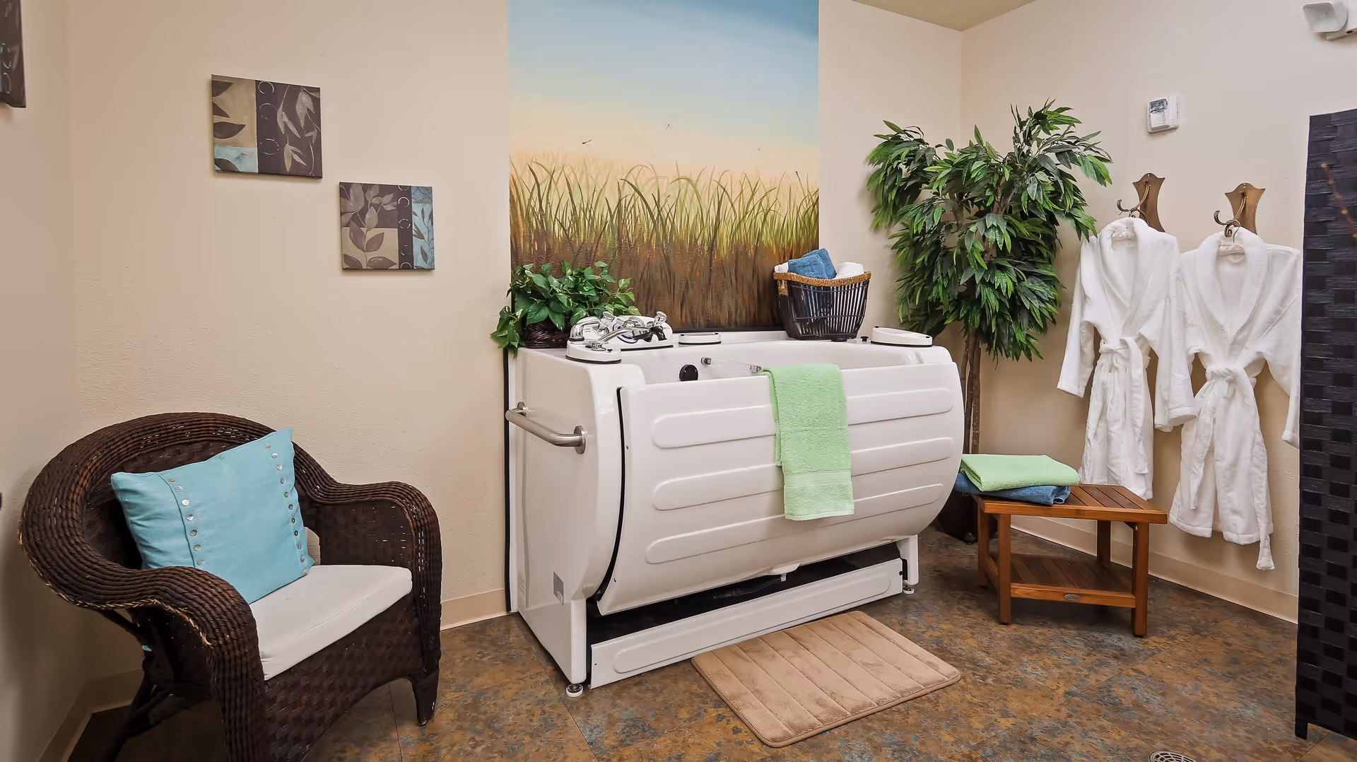 A cozy bathing room featuring a white walk-in bathtub with a green towel hanging on its side. Next to the tub is a wooden bench with folded towels and two white bathrobes hanging on wall hooks. A wicker chair with a light blue cushion is positioned to the left, and a large potted plant adds greenery to the corner. The wall behind the tub has a mural of tall grass under a blue sky, creating a calming atmosphere.
