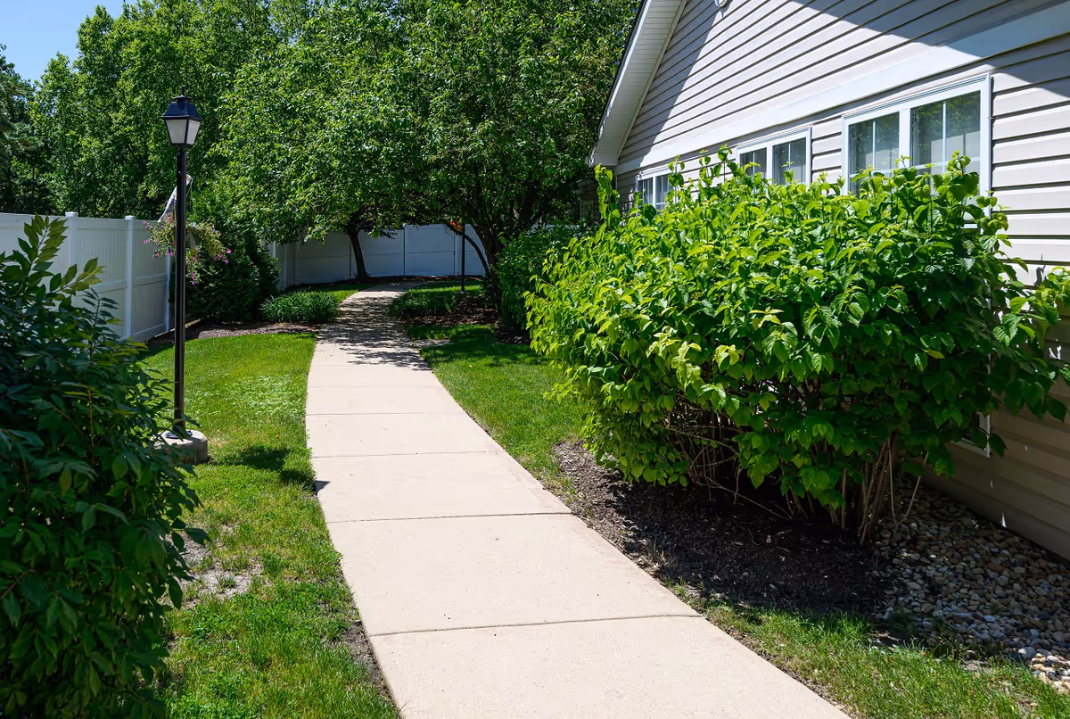 A paved walkway curves through a garden area with green grass, bushes, and trees. A beige building with white-framed windows is on the right side, and a white fence runs along the left side. A black lamp post with hanging flowers is visible near the walkway.