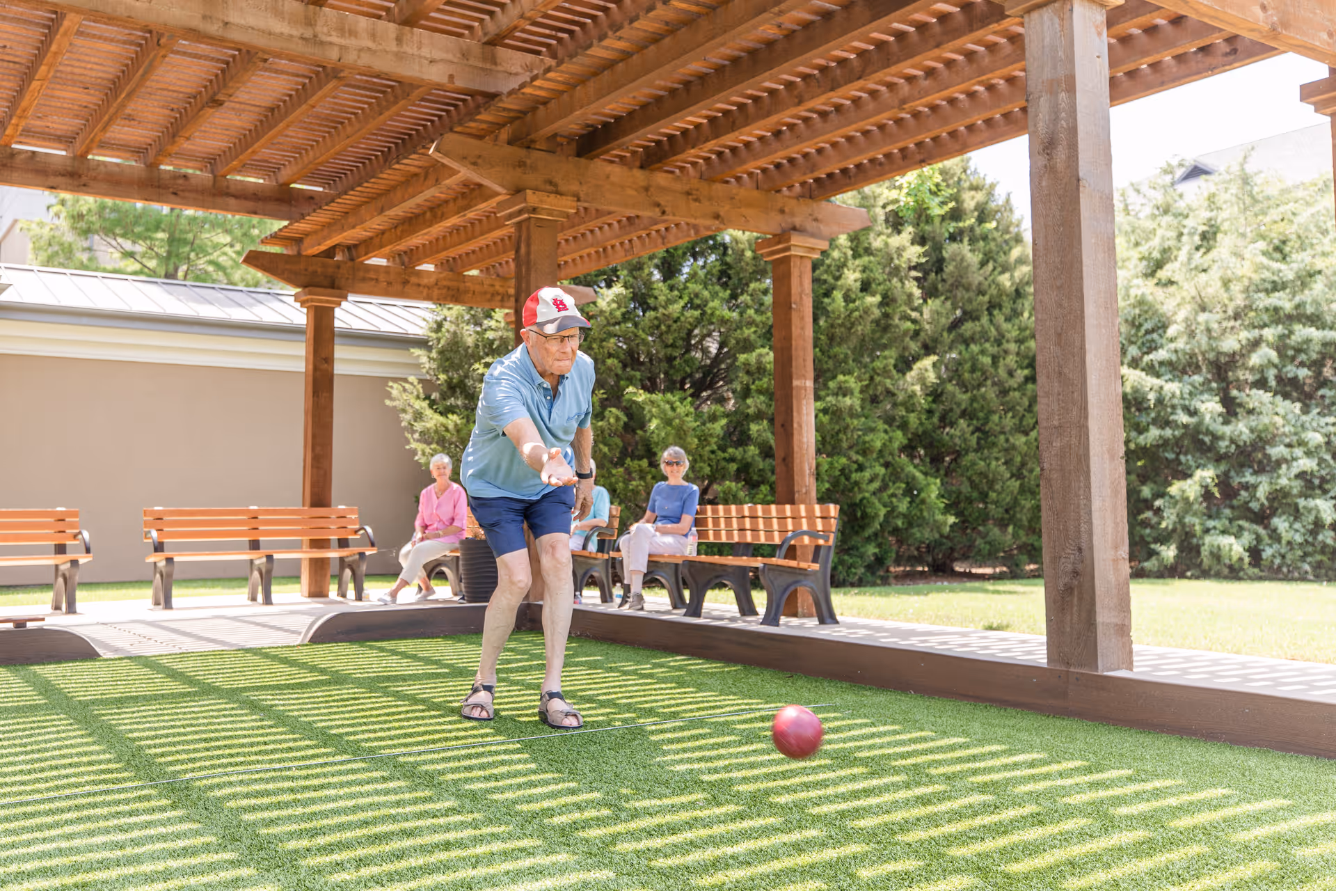 An elderly man wearing a red and white cap, blue shirt, and shorts is playing bocce ball on a green lawn under a wooden pergola. Two elderly women are sitting on benches in the background, watching the game. The area is surrounded by trees and greenery.