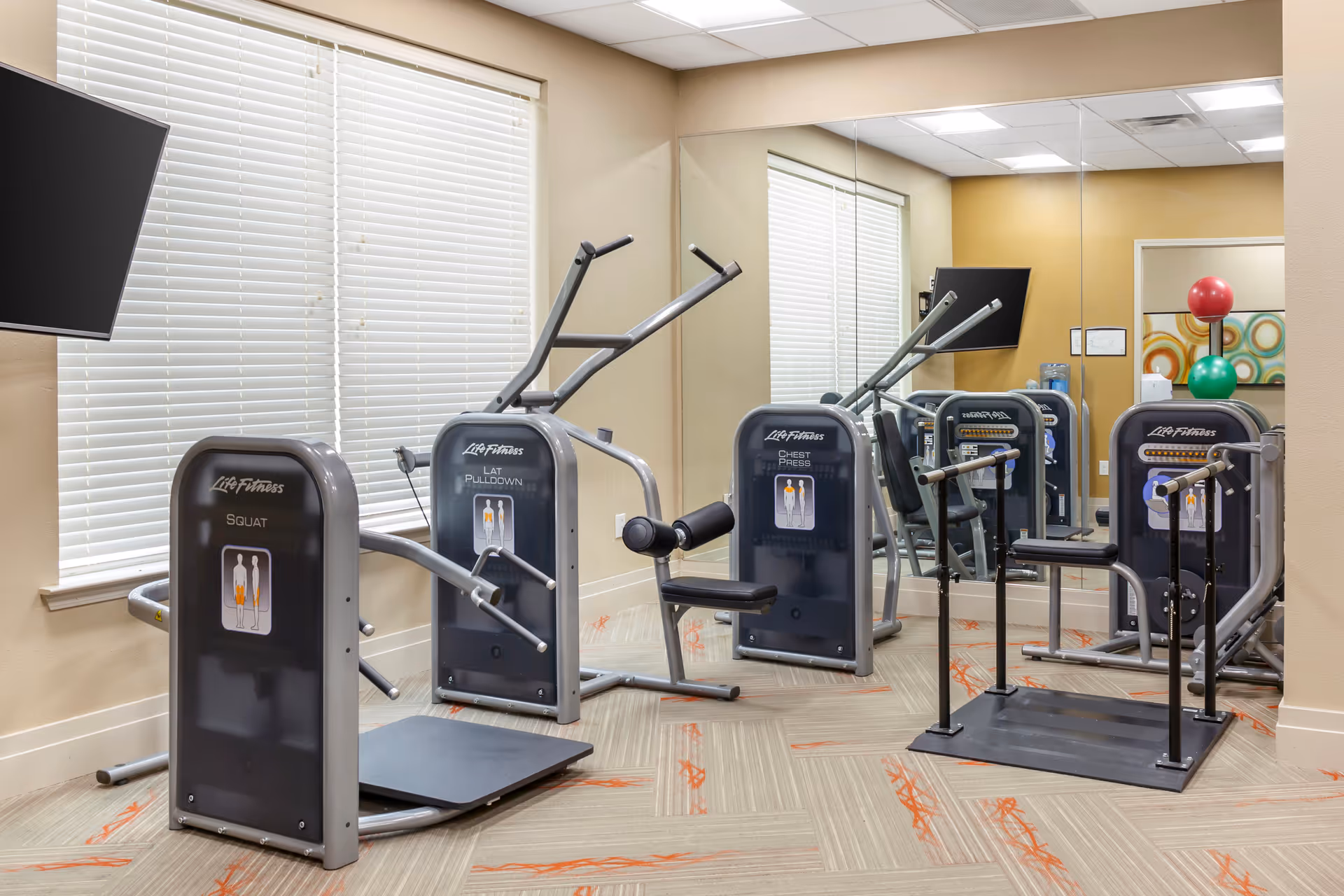A small fitness room with exercise machines including squat, lat pulldown, and chest press equipment. The room has beige walls, a large mirror on one side, a window with white blinds, a wall-mounted TV, and colorful exercise balls stacked in the corner.