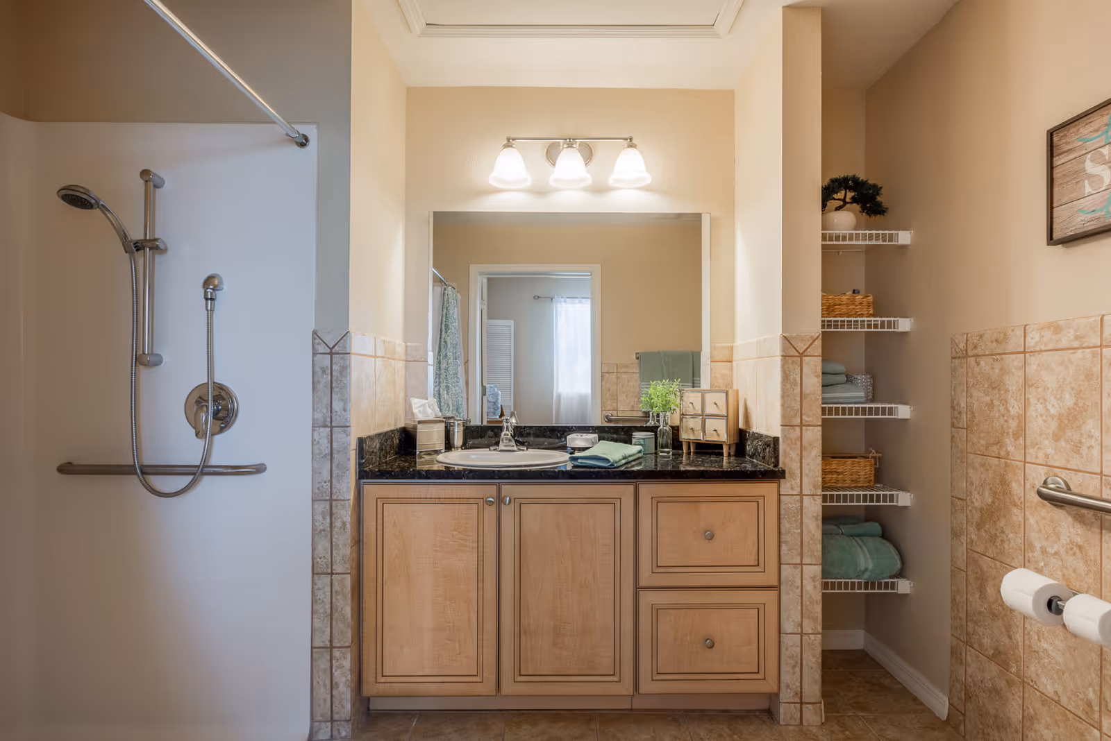 A bathroom with a shower on the left side featuring a handheld showerhead and grab bar. In the center, there is a wooden vanity with a black countertop, a sink, and a large mirror above it. The vanity has two drawers and two cabinet doors. Above the mirror, there is a three-light fixture. On the right side, there are white wire shelves holding towels and baskets, and a wall-mounted grab bar with two toilet paper rolls. The walls are partially tiled with beige tiles.