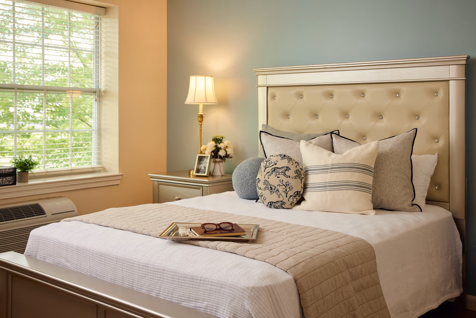 A neatly made bed with a tufted beige headboard, decorated with multiple pillows in various patterns and colors. A beige quilted throw is folded at the foot of the bed. Next to the bed is a nightstand with a lamp, a small framed photo, and a vase of flowers. A window with blinds lets in natural light, and a small potted plant sits on the windowsill.