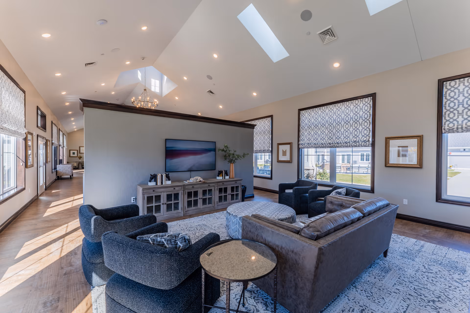 A spacious and well-lit living room area in a senior living facility featuring a large brown leather sofa, three dark upholstered armchairs, a round ottoman, and a wooden TV console with a flat-screen television mounted above it. The room has large windows with patterned blinds, framed artwork on the walls, skylights in the ceiling, and a chandelier. Hardwood floors and a patterned area rug complete the cozy and inviting atmosphere.