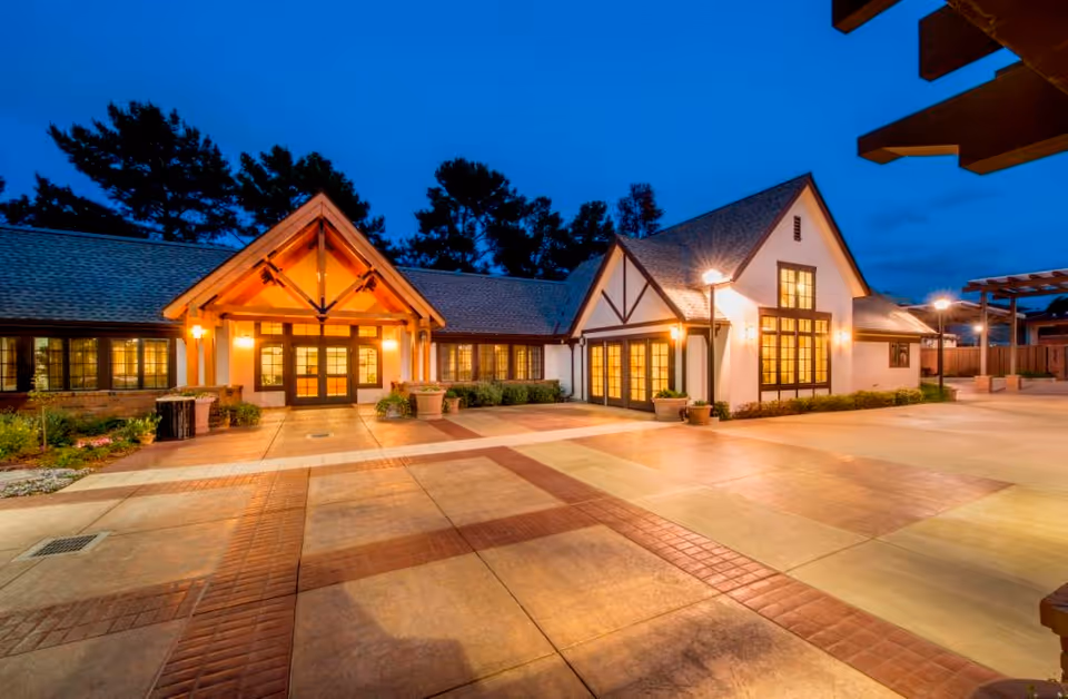 Exterior view of a senior living community building at dusk with warm lights glowing from the windows and entrance. The building features a pitched roof and decorative wooden beams, with a spacious paved courtyard in front.
