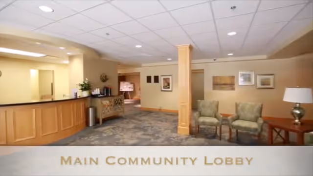 A spacious senior living community lobby with a wooden reception desk on the left, a decorative cabinet, two green upholstered chairs, a wooden side table with a lamp, and framed artwork on the beige walls. The floor is carpeted with a patterned design and the ceiling has recessed lighting.