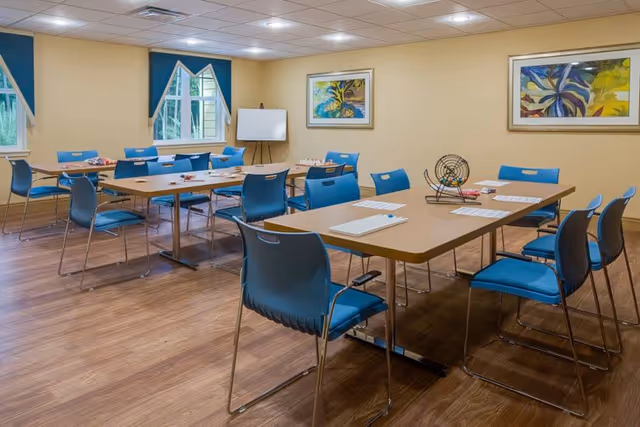 A well-lit room with several tables and blue chairs arranged for group activities. The tables have bingo cards and a bingo cage, indicating a recreational or game setting. The room has wooden flooring, beige walls, two framed colorful paintings, and windows with blue curtains.