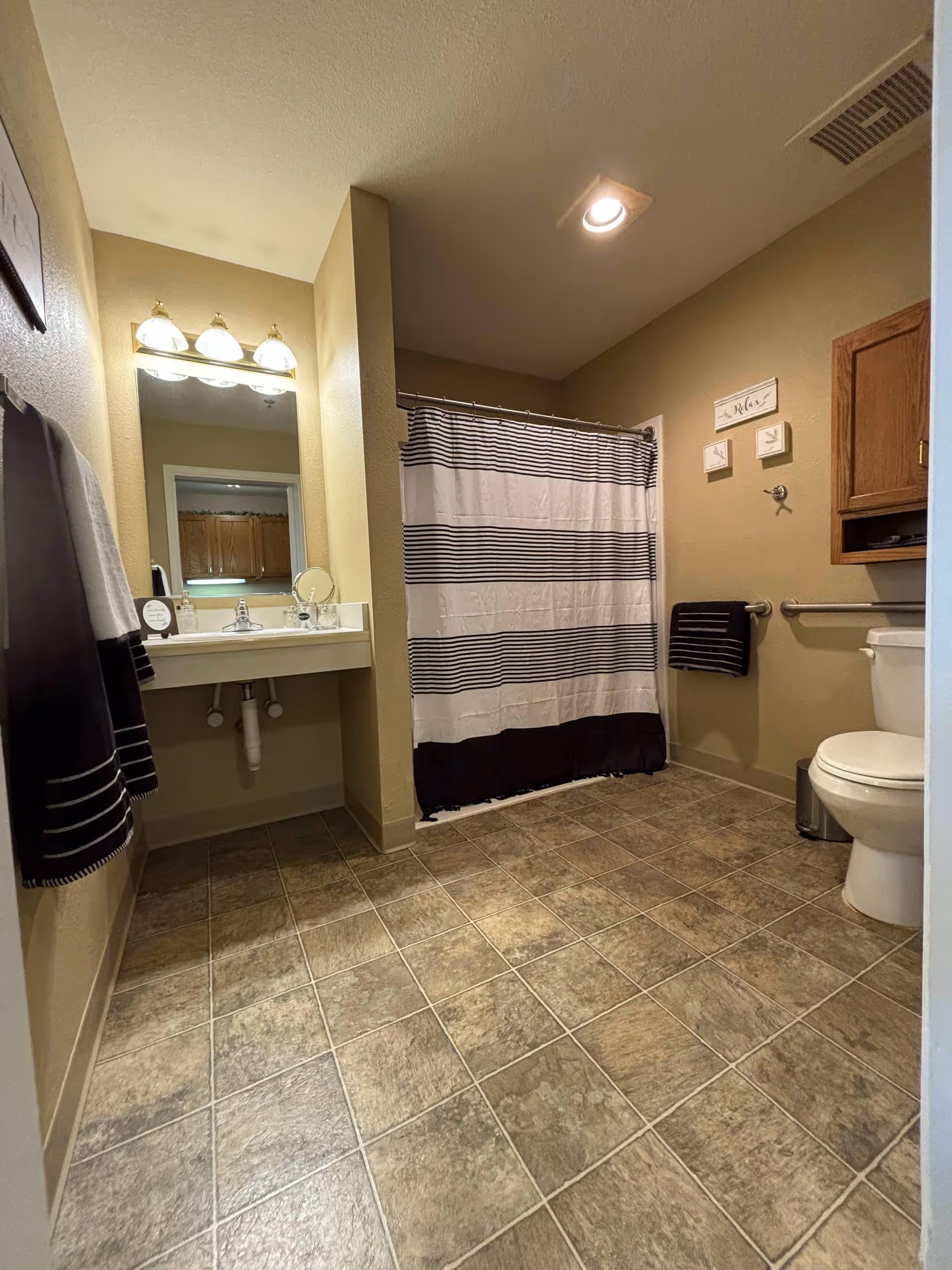 A bathroom with beige walls and tiled floor. There is a white sink with a mirror and three light fixtures above it on the left side. A towel rack with black and white towels is mounted on the wall near the sink. In the back, there is a bathtub with a white and black striped shower curtain. On the right side, there is a toilet with a wooden cabinet above it and a small trash can next to it. The bathroom has a clean and organized appearance.