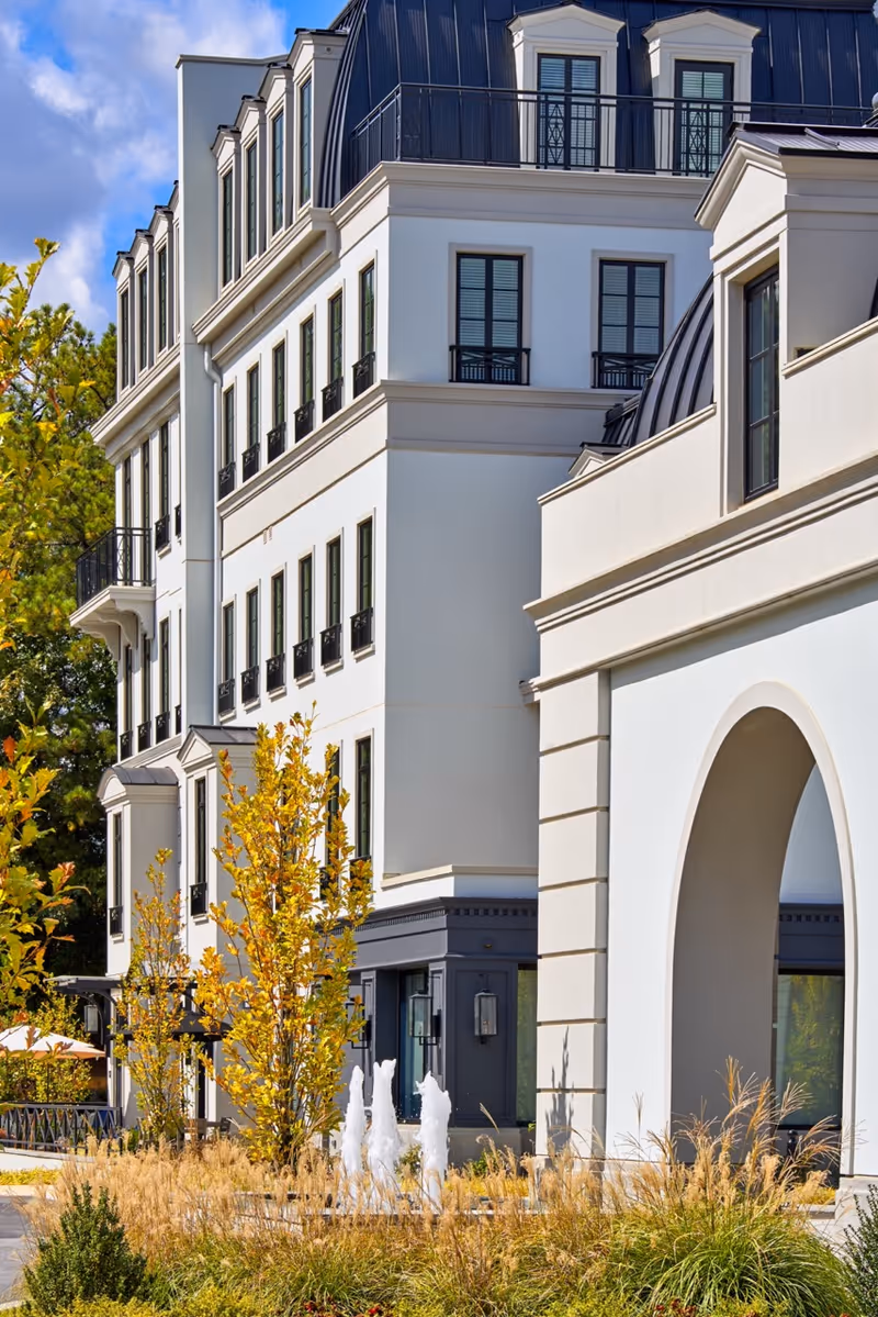 Exterior view of a multi-story building with white walls and black window frames, featuring a landscaped area with ornamental grasses, small trees with yellow leaves, and a water fountain in front. The sky is blue with some clouds.