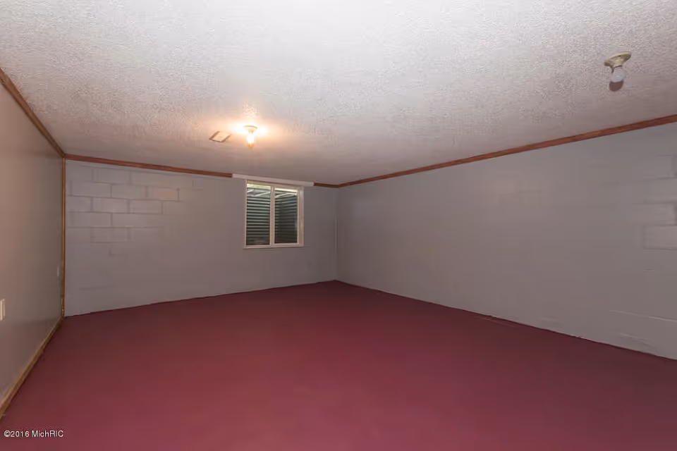 Empty room with gray painted cinder block walls, a small window with an exterior window well, a maroon carpeted floor, and a textured white ceiling with two exposed light bulbs.