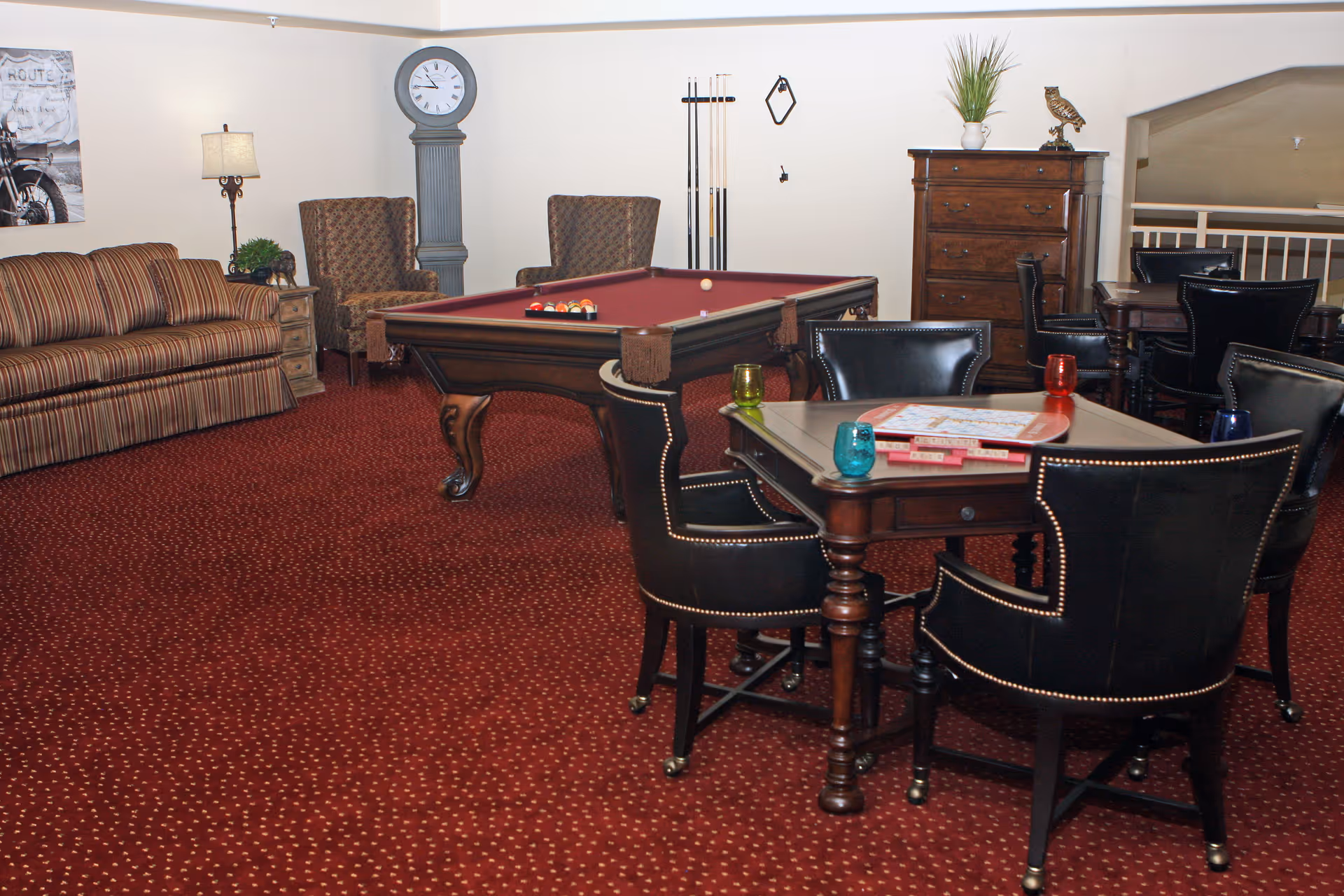 A cozy game room with a red carpet featuring small yellow dots, a pool table with balls set up, a table with board games and four black leather chairs, a striped sofa, two patterned armchairs, a tall grandfather clock, a wooden chest of drawers with a plant and owl figurine on top, and a floor lamp next to the sofa.