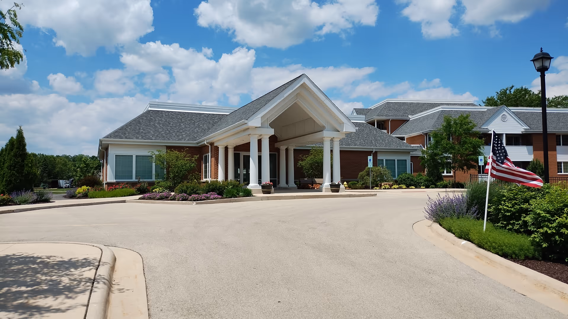 Exterior view of Peterson Meadows senior living facility showing a large brick building with white columns at the entrance, surrounded by well-maintained landscaping, an American flag, and a clear blue sky with some clouds.