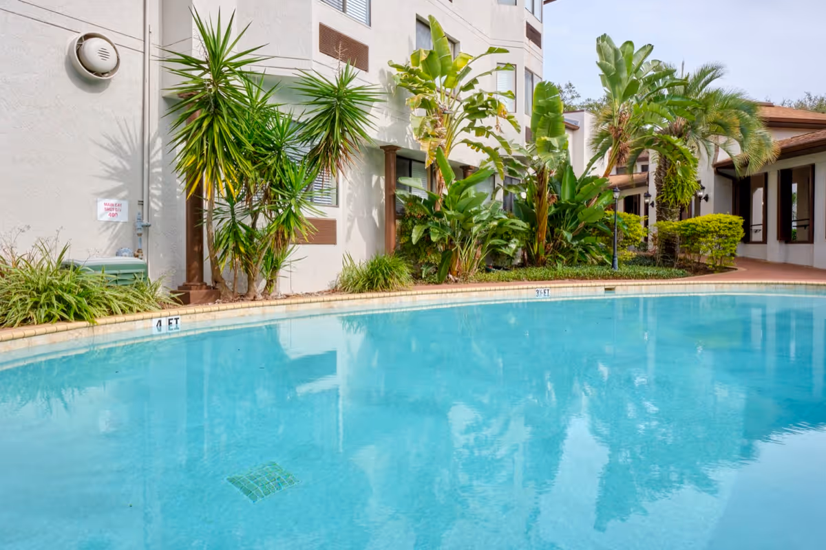 Outdoor swimming pool surrounded by tropical plants and trees, adjacent to a multi-story building with windows and a covered walkway.