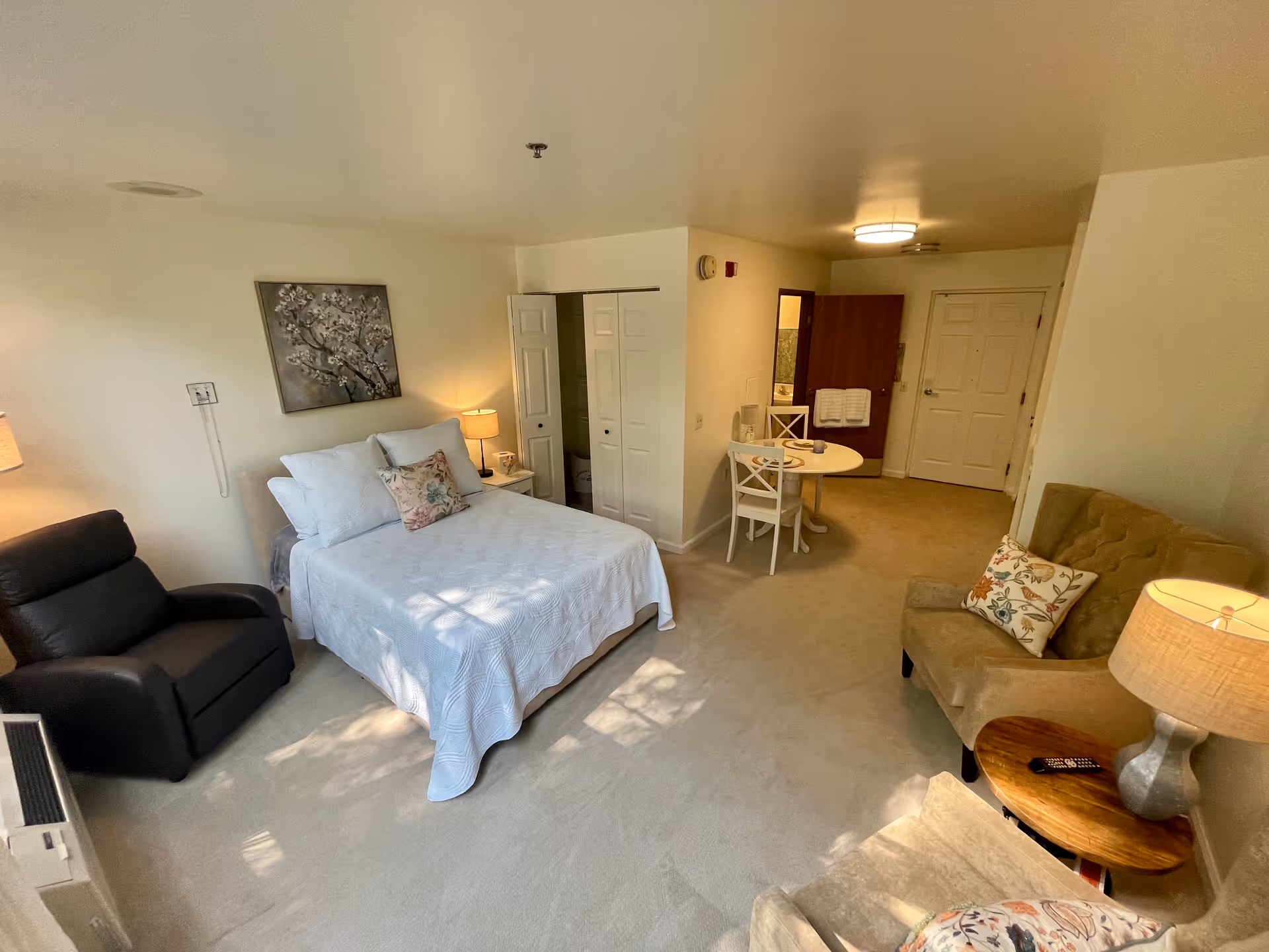 A cozy senior living facility room with a bed covered in white bedding and a floral pillow, a black recliner chair to the left, and a beige armchair with a floral pillow to the right. There is a small round table with two chairs near the entrance door. The room has beige carpet and walls, a painting above the bed, and a lamp on a side table next to the armchair.