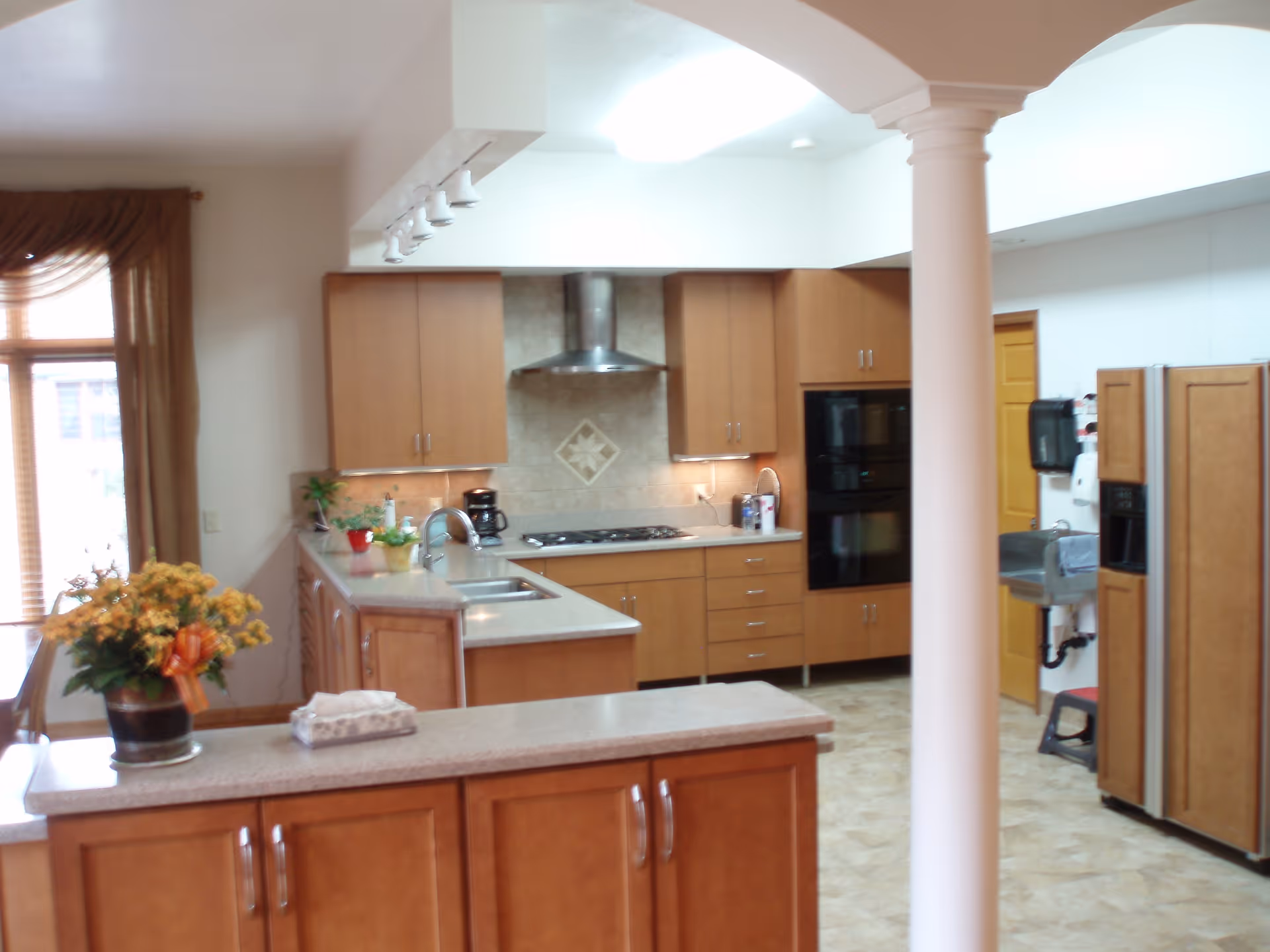 A spacious kitchen with wooden cabinets and beige countertops. The kitchen features a double oven, a gas stove with a stainless steel range hood, a double sink, and a side-by-side refrigerator. There is a small potted plant and a coffee maker on the counter. A large window with brown curtains allows natural light into the room. A white column stands in the foreground.