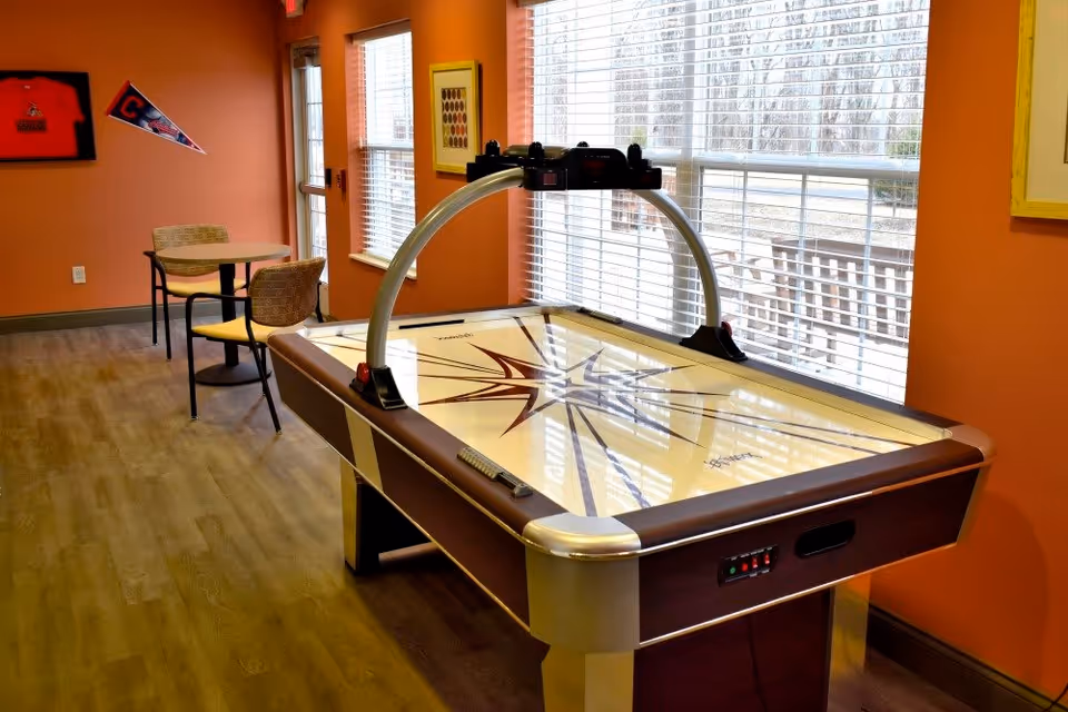 A game room with an air hockey table in the foreground and a small round table with two chairs near a door and windows with blinds. The walls are painted orange and decorated with a framed sports jersey and a pennant.