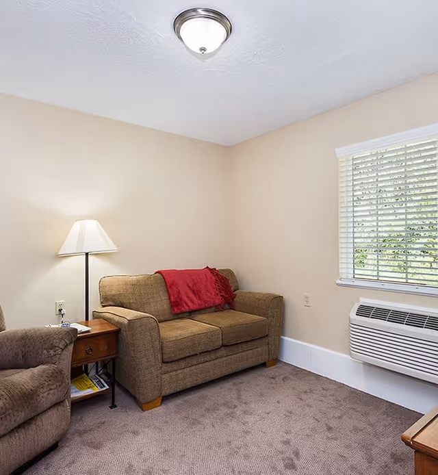 Small living room with a loveseat and armchair, side table with lamp, window blinds, and a wall air-conditioning unit.