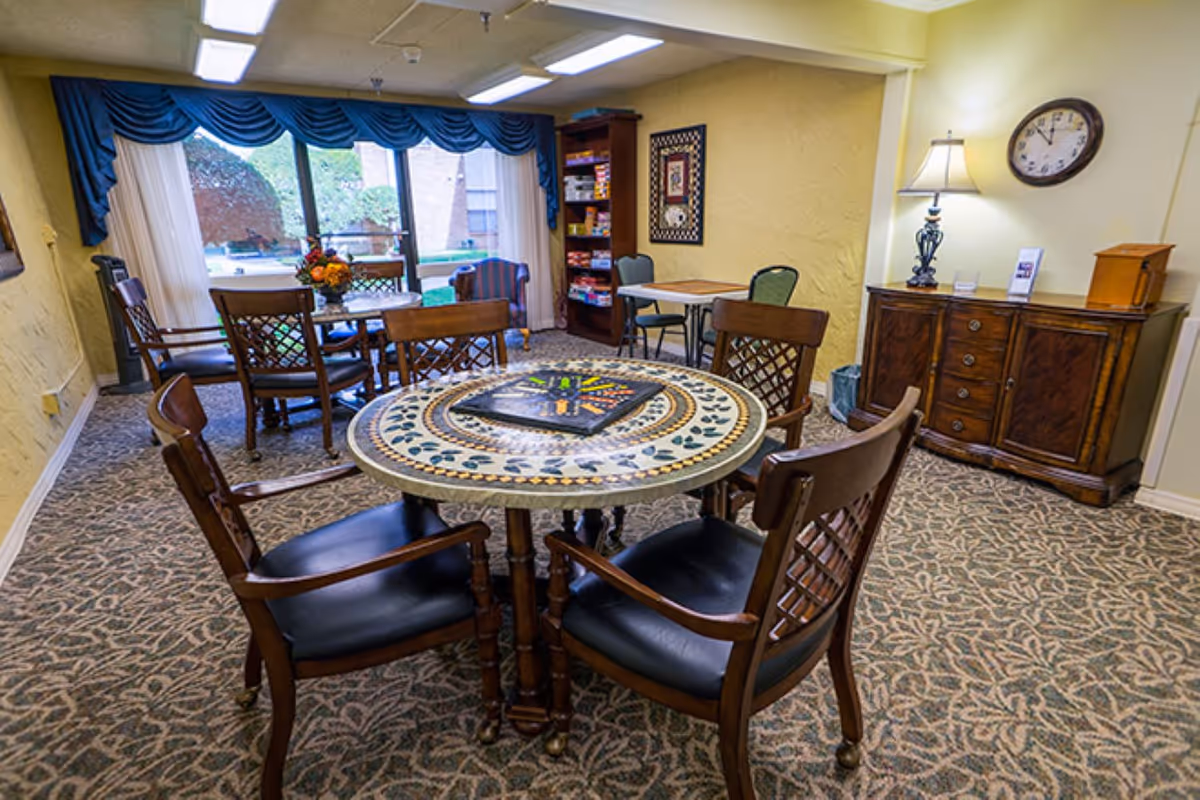 A cozy common area with a round mosaic table surrounded by four wooden chairs with black cushions. In the background, there is another table with chairs, a tall bookshelf filled with board games, a wooden cabinet with a lamp and clock on the wall above it, and large windows with blue valances letting in natural light.