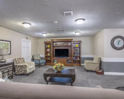 A cozy senior living common area with a large wooden entertainment center holding a flat-screen TV and decorative items. The room has several upholstered chairs and a sofa arranged around a dark wooden coffee table with a floral centerpiece. The walls are painted in neutral tones with white trim, and a round clock is mounted on the wall to the right.