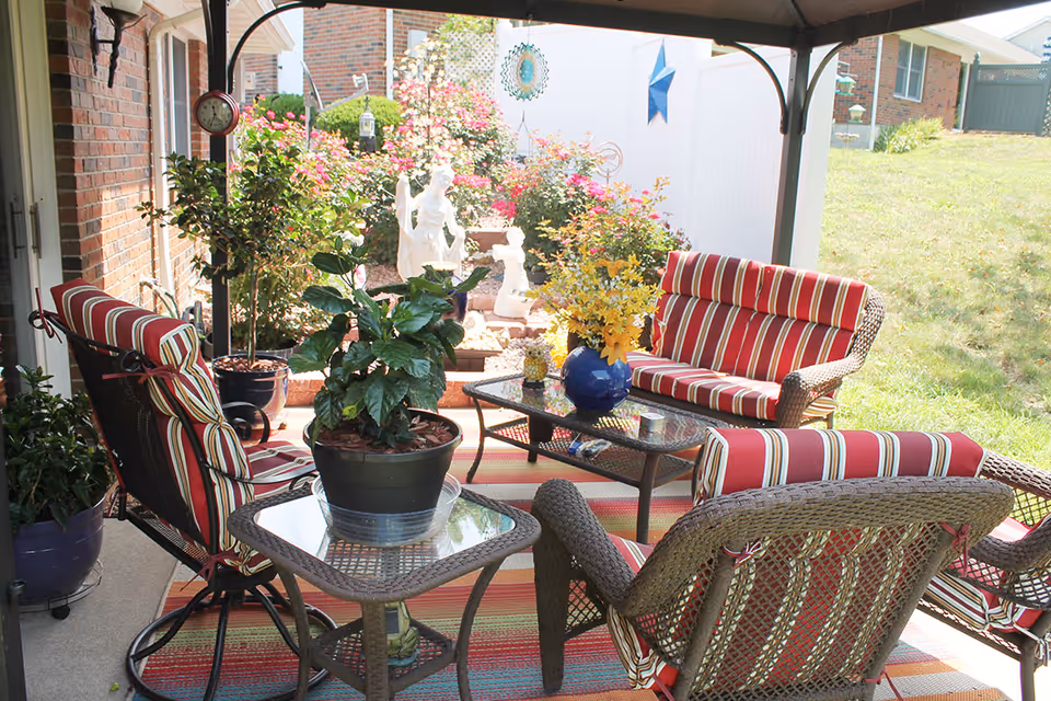 Covered outdoor patio area with wicker furniture including chairs and a loveseat with red and white striped cushions. There are several potted plants on glass-top tables and a colorful striped rug on the floor. In the background, there is a garden with blooming flowers, decorative statues, and a white fence.