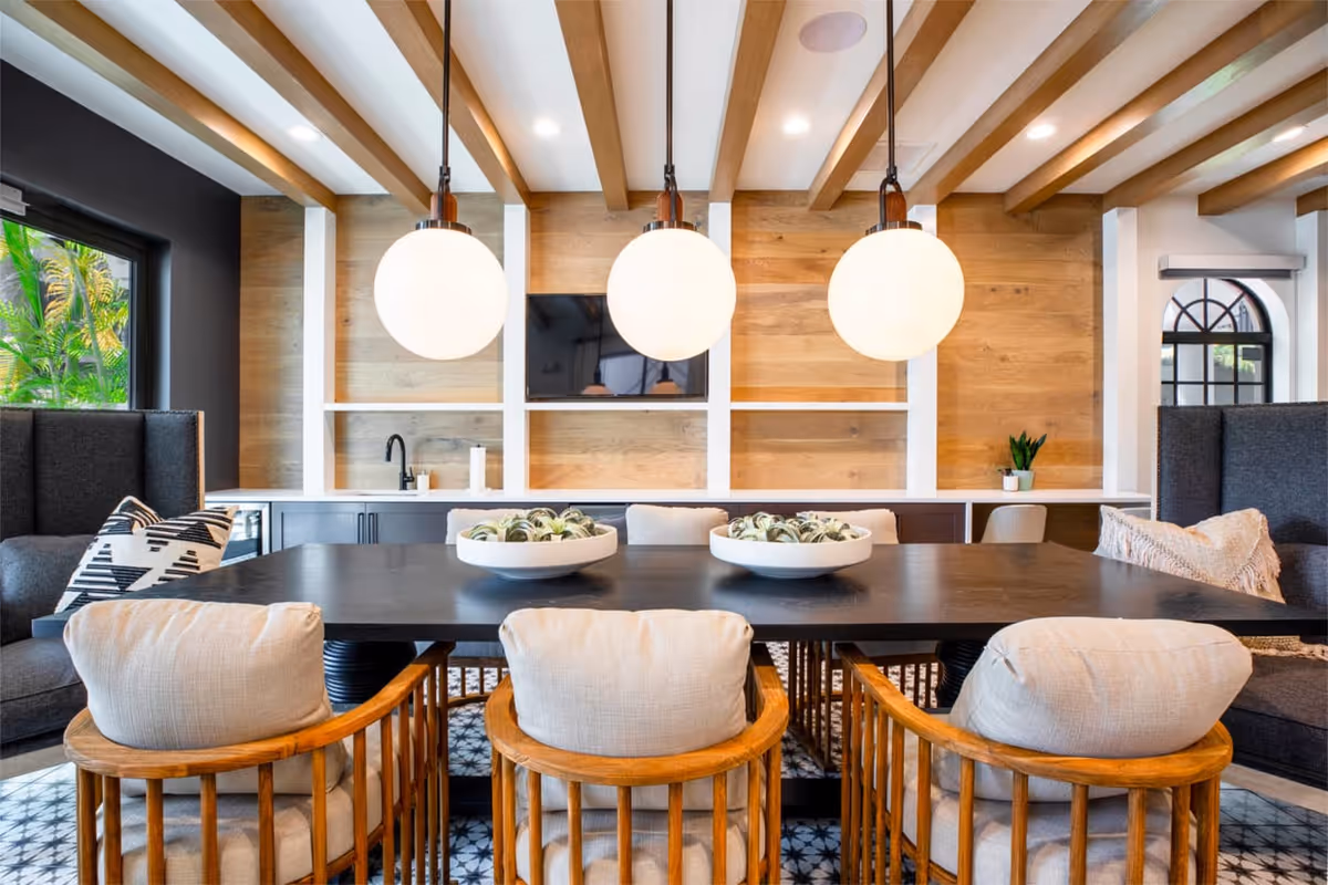 A modern dining area with a large black table surrounded by cushioned wooden chairs and two high-backed gray armchairs. Three round pendant lights hang from a ceiling with wooden beams. The back wall features wooden paneling with white shelving, a mounted flat-screen TV, and a countertop with a sink and small plants. A window on the left shows greenery outside.