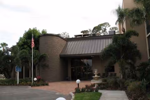 Exterior view of River Commons facility entrance with curved tower, flag, and landscaped walkway.