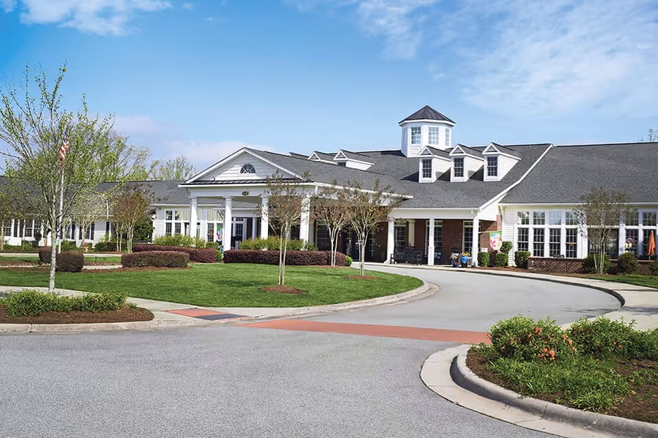 Front exterior view of a large, single-story senior living facility building with white siding, multiple windows, a covered entrance with columns, and a circular driveway surrounded by well-maintained landscaping and small trees under a partly cloudy sky.