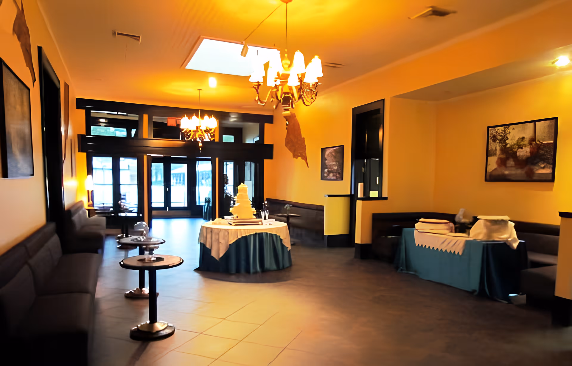 Interior view of a common area with yellow walls and dark wood trim, featuring multiple seating areas with cushioned benches and small round tables. A round table in the center is covered with a white and blue tablecloth and holds a multi-tiered white cake. Another table on the right side is covered with a blue tablecloth and has folded linens or towels on top. The room is illuminated by chandeliers and natural light from large windows and glass doors at the far end.