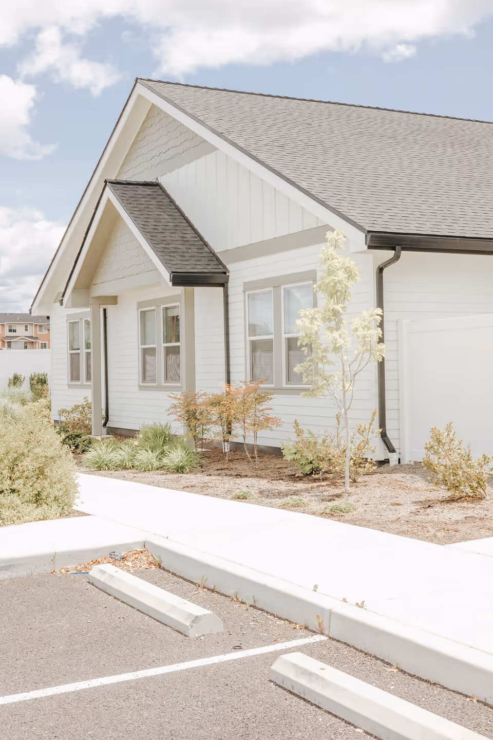 Exterior view of a single-story white building with a gray shingled roof, several windows, small shrubs, and a tree planted along the side. A parking area with marked spaces is visible in the foreground under a partly cloudy sky.