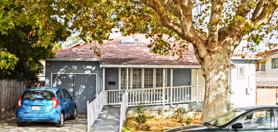 Front exterior view of a single-story residential building with a sloped roof, a large tree in the front yard, a white railing ramp leading to the entrance, and two parked cars in the driveway.