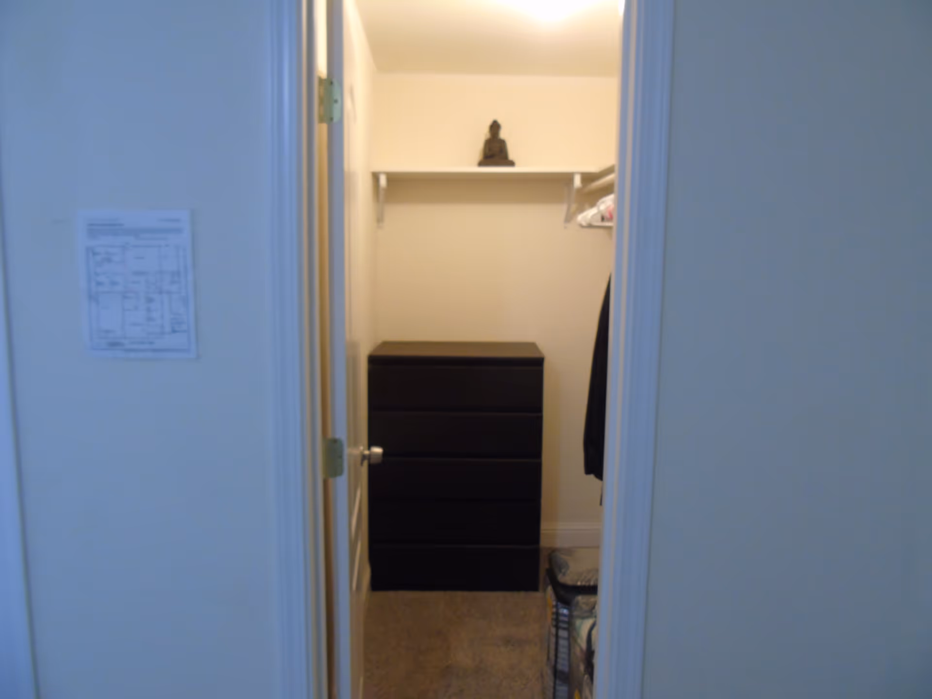 View through an open doorway into a small closet with a black chest of drawers, a shelf with a small Buddha statue on top, and some hanging clothes on the right side. A floor plan is posted on the wall outside the closet.