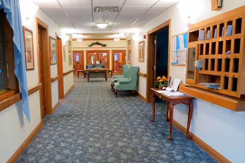 A well-lit hallway in a senior living facility with patterned carpet, wooden trim, framed pictures on the walls, a green armchair, a small wooden table with papers and a flower arrangement, and a wooden mail organizer. At the end of the hallway, there are glass doors leading to another room with more seating.