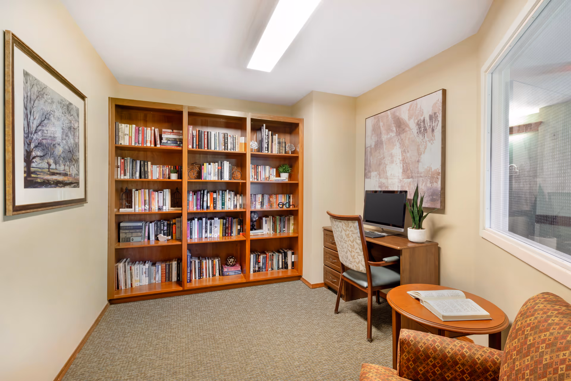 A cozy reading and computer room with a large wooden bookshelf filled with books, a wooden desk with a computer and a potted plant, a patterned armchair, and a small round table with an open book. The walls are beige with framed artwork, and there is a window with a view into another room.