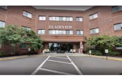 Front exterior view of Gladeview Health Care Center, a three-story brick building with large windows and a covered entrance. There are trees and shrubs on either side of the entrance and a driveway with marked pedestrian crossing lines in front.