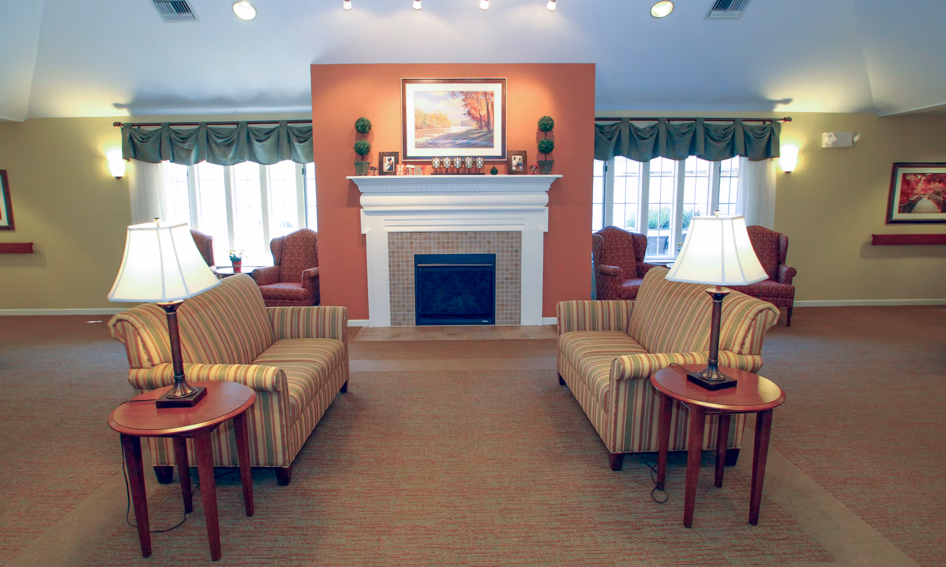 A cozy living room area with two striped sofas facing each other, each accompanied by a wooden side table with a lamp. In the background, there is a white fireplace with a reddish-brown accent wall, decorated with framed pictures and small plants. Large windows with green valances allow natural light to fill the room, and two upholstered armchairs are positioned near the windows.