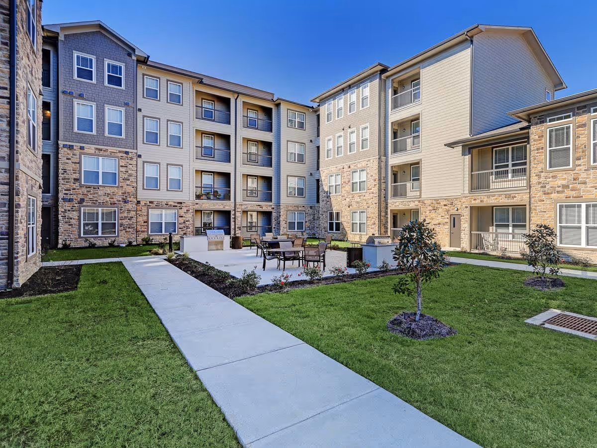 Outdoor courtyard area of a senior living facility with green grass, small trees, a concrete walkway, and a seating area with tables and chairs. The surrounding building has multiple floors with balconies and windows, featuring a combination of stone and siding exterior under a clear blue sky.