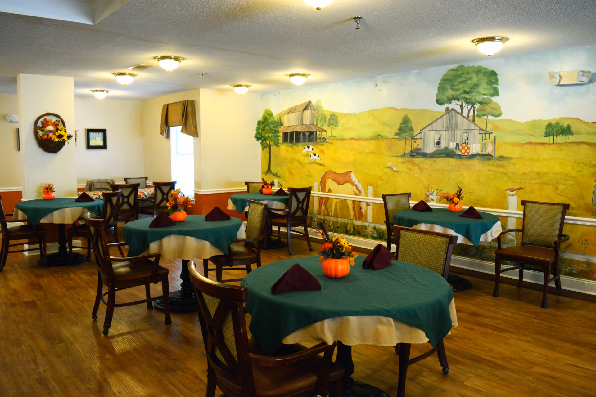 Dining room with round tables covered in green and beige tablecloths, each decorated with small orange pumpkin centerpieces and folded dark napkins. Wooden chairs surround the tables. The room has a wooden floor and a mural on the wall depicting a rural scene with a barn, horses, trees, and a farmhouse.
