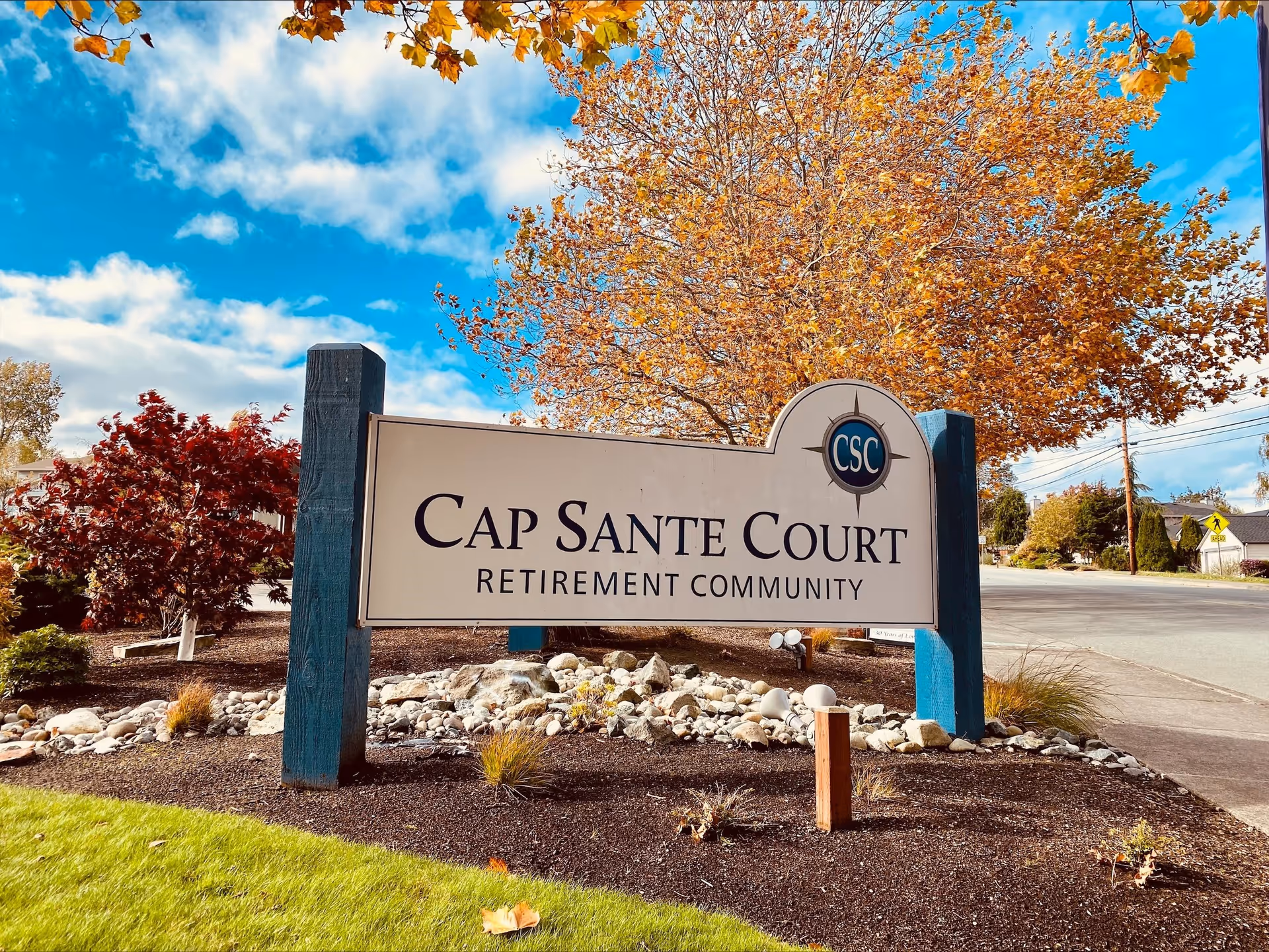 Outdoor view of a sign for Cap Sante Court Retirement Community surrounded by landscaped rocks, plants, and trees with autumn foliage under a partly cloudy blue sky.