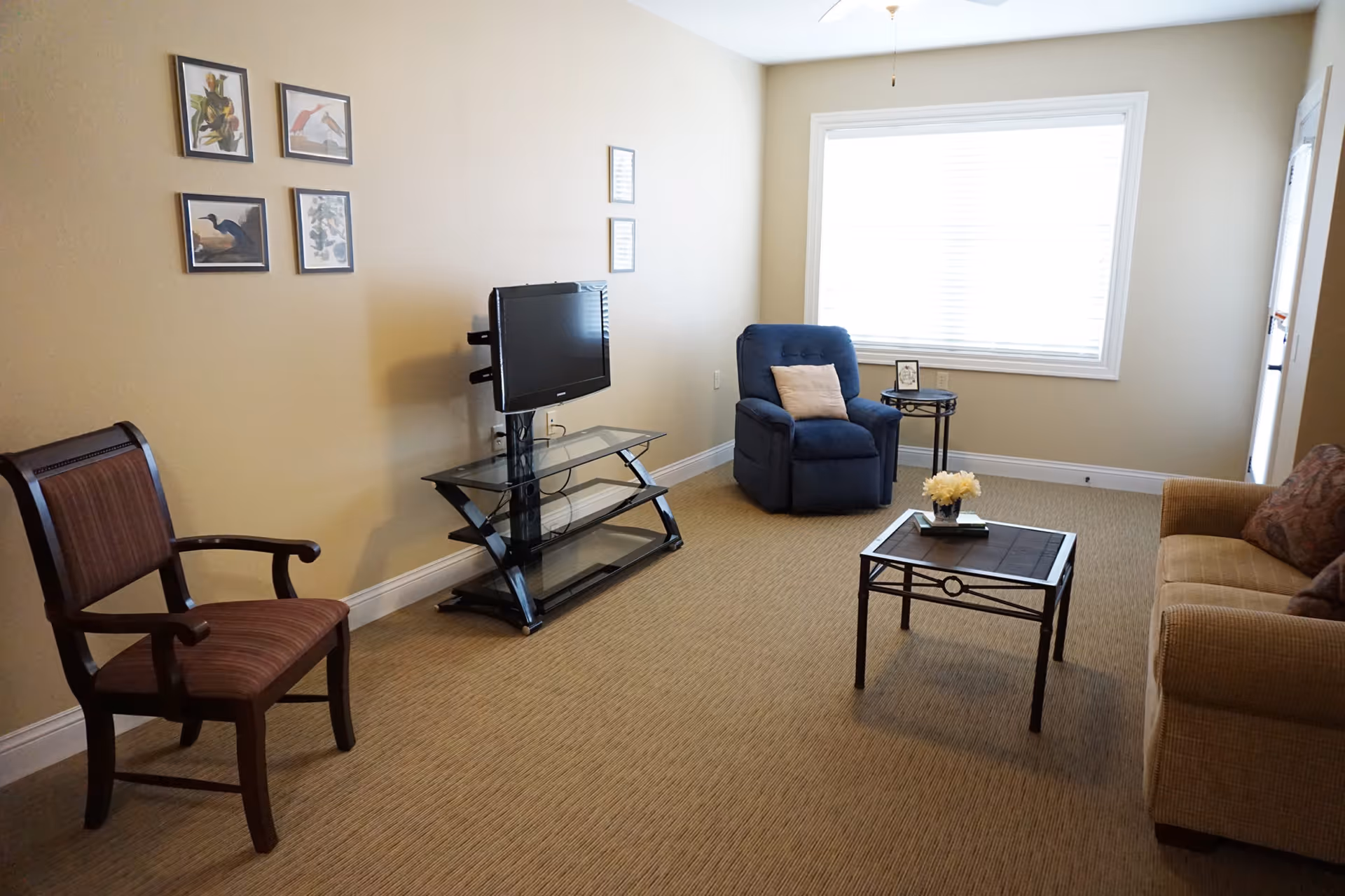 Simple living room with a TV on a glass stand, a blue recliner, sofa and chairs around a coffee table by a window.