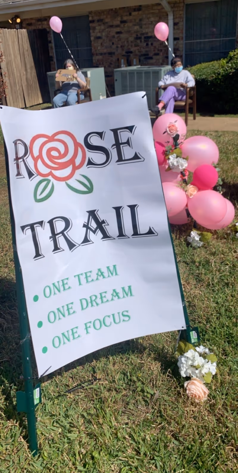 Outdoor scene at Avir at Rose Trail facility showing a white sign with a red rose graphic and the text 'ROSE TRAIL' along with the phrases 'ONE TEAM, ONE DREAM, ONE FOCUS'. Two people are seated in chairs in the background, each holding a pink balloon, with one person holding a sign that says 'I love you'. Pink and white balloons and flowers are arranged on the grass nearby.