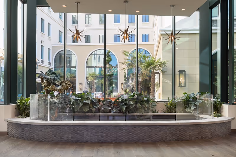 Indoor garden area with a curved stone planter filled with green plants, surrounded by glass panels. Large floor-to-ceiling windows behind the planter reveal an exterior courtyard with palm trees and a beige building with arched windows. Three modern star-shaped pendant lights hang from the ceiling above the planter.
