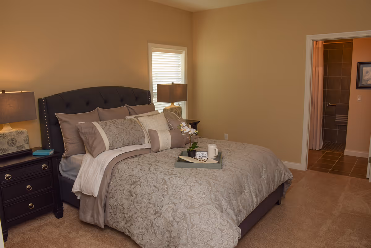 A neatly made bed with multiple pillows and a patterned comforter in a bedroom. There are two bedside tables with lamps on either side of the bed. A tray with a white mug, a small plant, and a pair of glasses rests on the bed. A window with blinds is visible behind the bed, and an open doorway leads to a bathroom with tiled walls and floor.