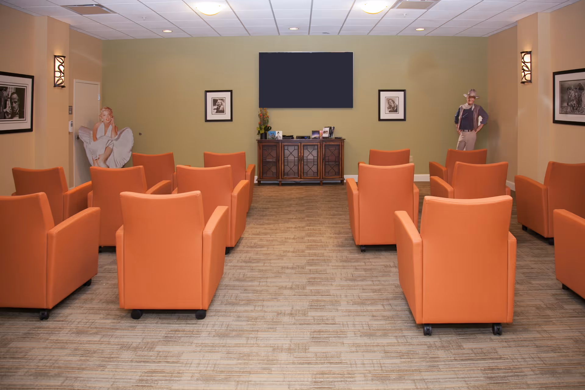 A small theater or media room with three rows of orange armchairs facing a large flat-screen TV mounted on a green wall. Below the TV is a wooden cabinet with decorative items. The walls are decorated with framed black and white photographs and two life-size cardboard cutouts of a woman and a man. The room has a carpeted floor and ceiling lights.