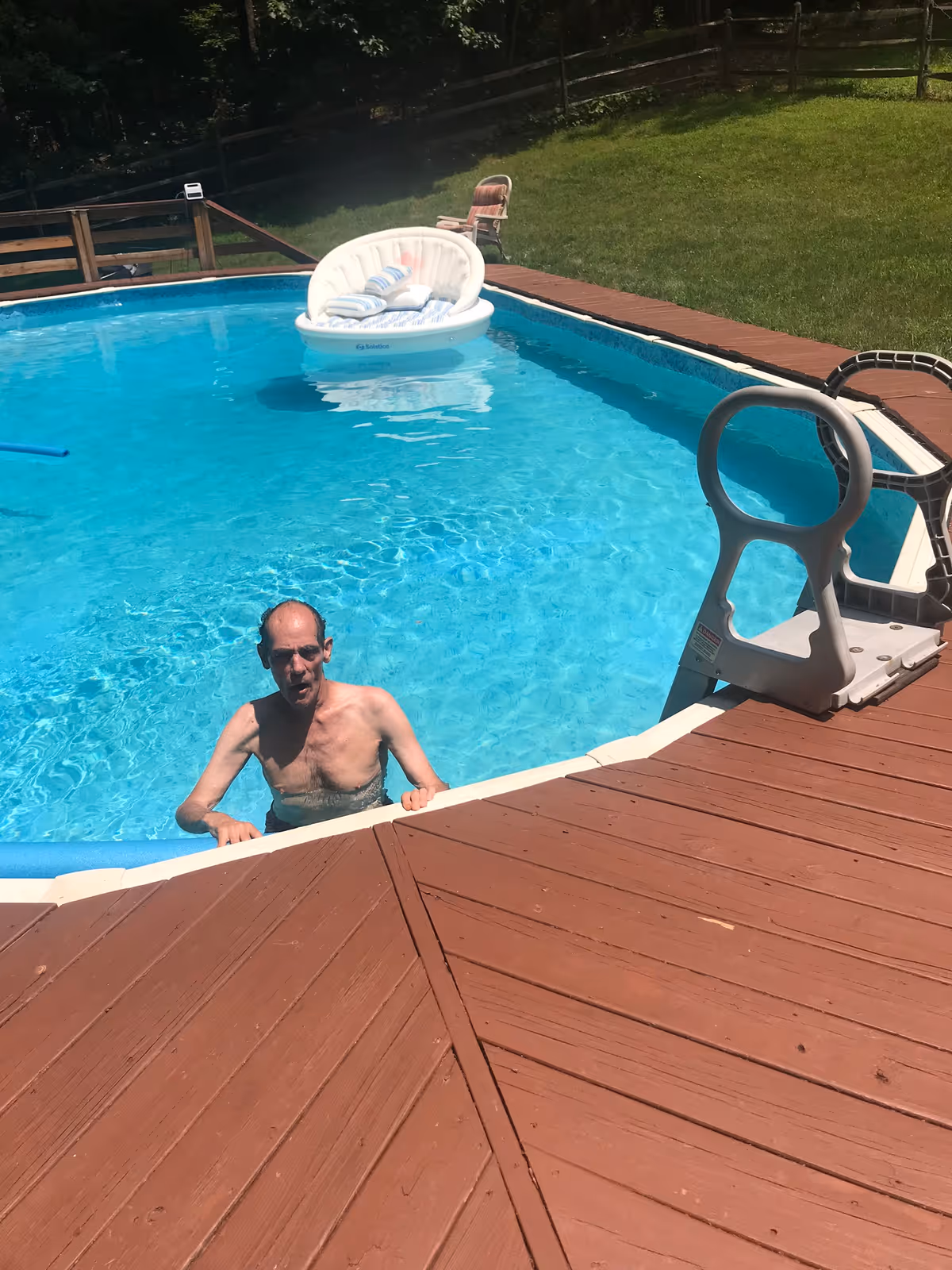 An elderly man is standing in an above-ground swimming pool with clear blue water. The pool is surrounded by a wooden deck, and there is a white inflatable pool float with pillows on the water. In the background, there is a grassy area with a wooden fence and a chair.