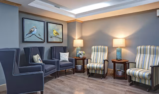 A cozy sitting area with six armchairs arranged along the walls. Three dark blue armchairs with decorative pillows are on the left side, and three striped armchairs in light colors are on the right. Two wooden side tables with lamps and a small flower arrangement are placed between the chairs. Two framed bird paintings hang on the wall above the blue chairs. The room has wood flooring and soft lighting.