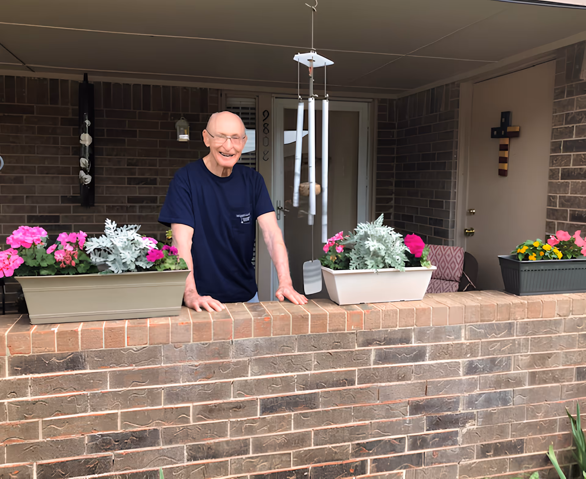 An elderly man wearing glasses and a navy blue t-shirt stands behind a brick patio wall with flower pots containing pink and yellow flowers. Behind him is a brick wall with a door, a window, a hanging wind chime, and a decorative cross on the wall.