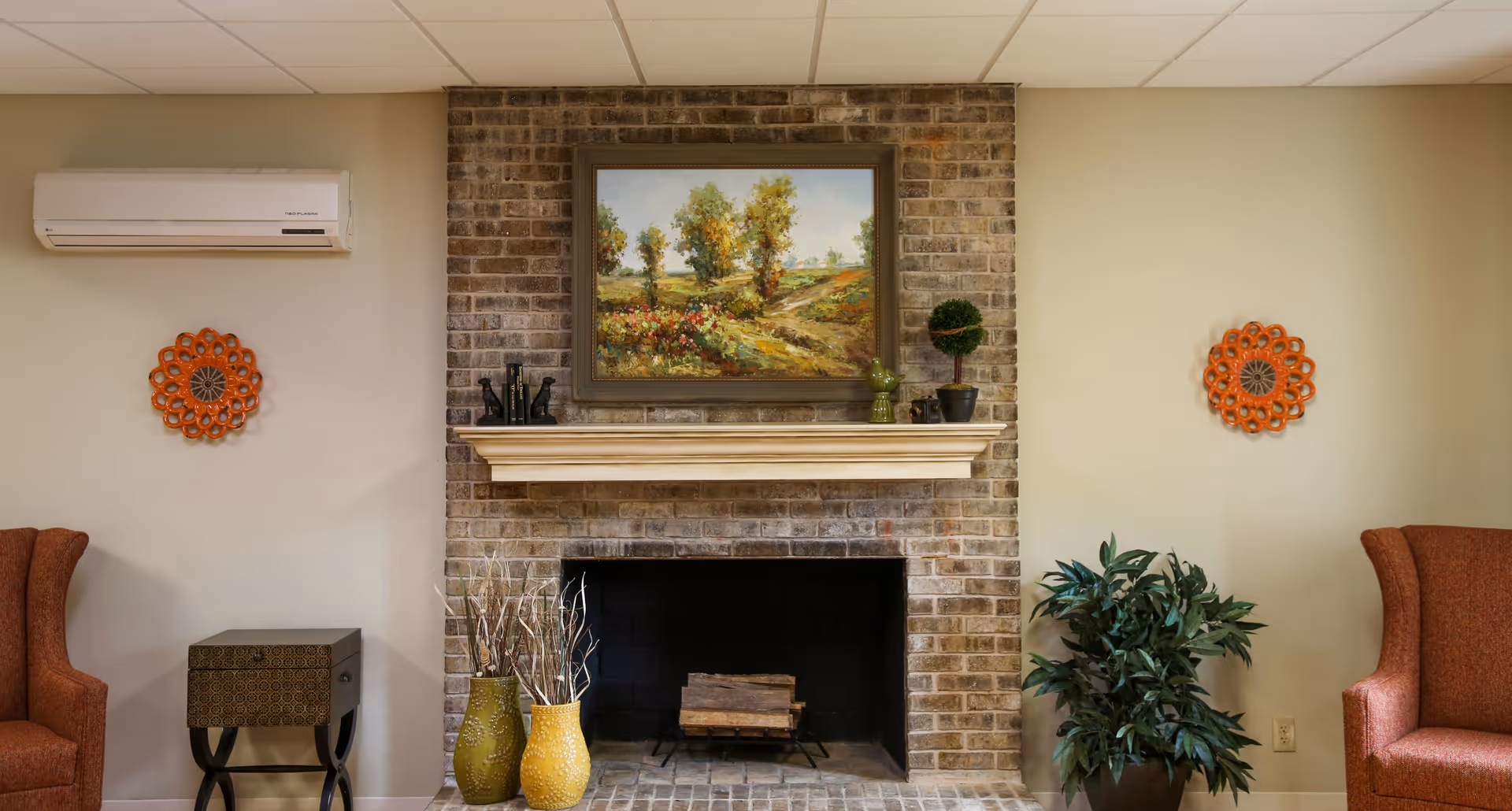 A cozy living room area with a brick fireplace in the center. Above the fireplace is a framed painting of a landscape with trees and flowers. On the mantel are decorative items including small plants and figurines. On either side of the fireplace are beige walls with orange decorative wall hangings. There are two orange upholstered armchairs, one on each side, a small side table on the left, and a green potted plant on the right. The ceiling has white tiles and a wall-mounted air conditioning unit is visible on the left wall.