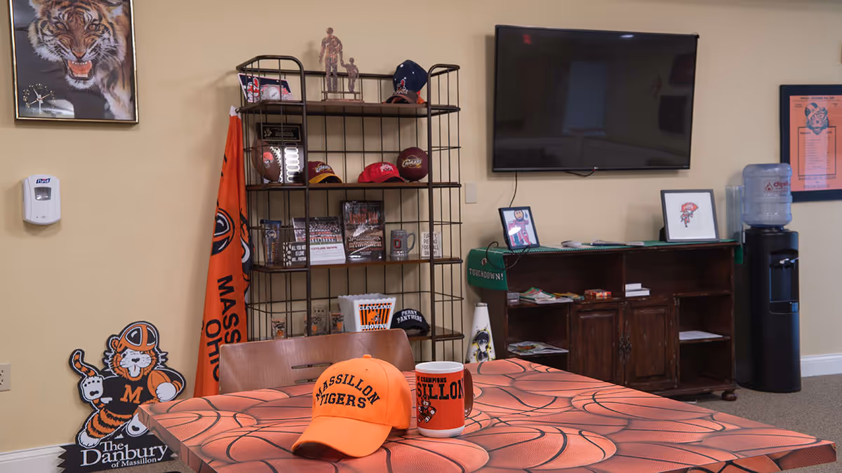 A room with a table covered in a basketball-themed tablecloth, an orange Massillon Tigers cap, and a matching mug. Behind the table is a metal shelving unit with sports memorabilia including hats, footballs, and framed photos. A large flat-screen TV is mounted on the wall above a wooden cabinet with more framed pictures and a water dispenser to the right. A tiger mascot poster and an orange flag with 'Massillon Ohio' are also visible on the left side of the image.