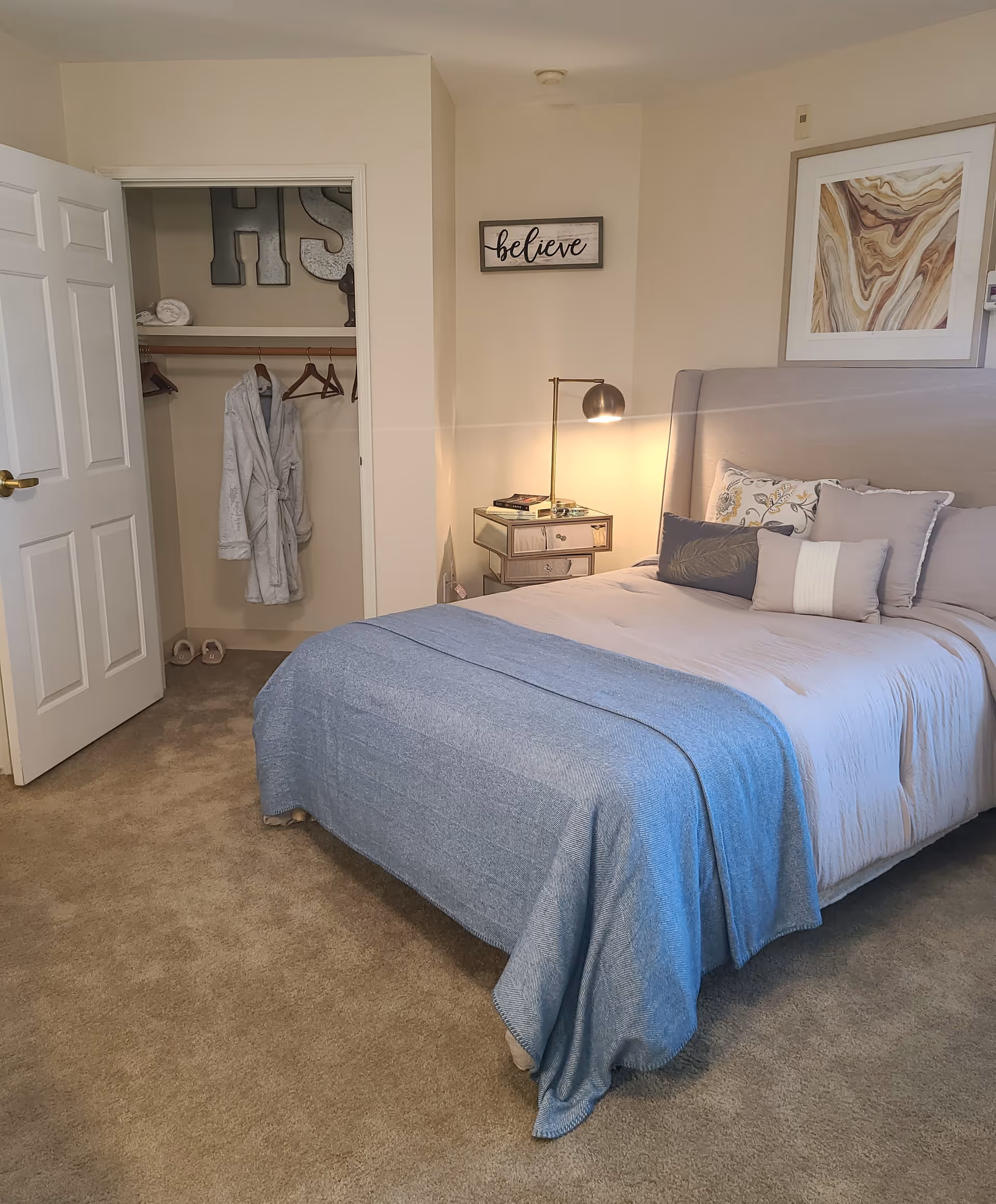 A cozy bedroom with a neatly made bed featuring multiple pillows and a blue throw blanket. To the right of the bed is a mirrored nightstand with a lamp and some books. On the wall above the nightstand is a framed abstract artwork and a small sign that says 'believe'. To the left, an open closet door reveals a hanging white robe and some hangers. The room has beige carpet and light-colored walls.